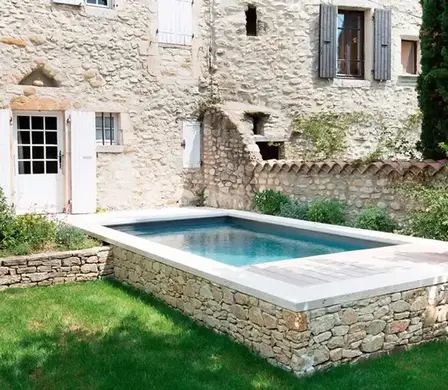Rechteckiger Pool in einer Natursteinmauer eingefasst. Rustikale Hausmauern im Hintergrund mit Garten.